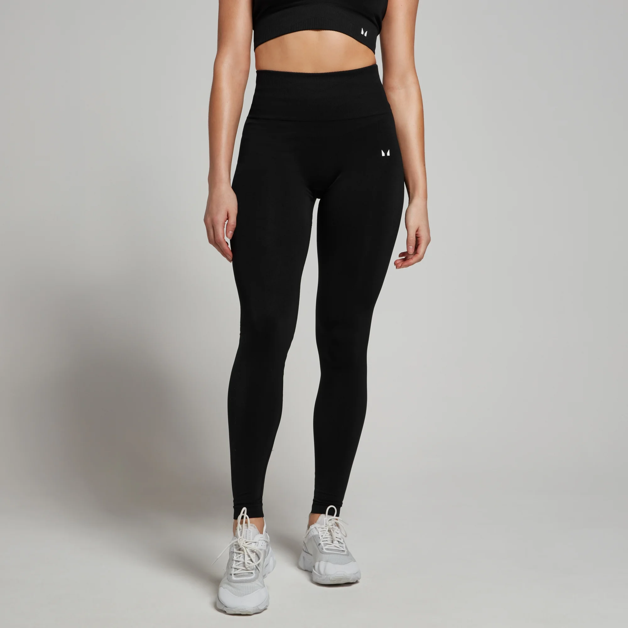 Een vrouw draagt een zwarte, naadloze legging met hoge taille en witte sportschoenen.