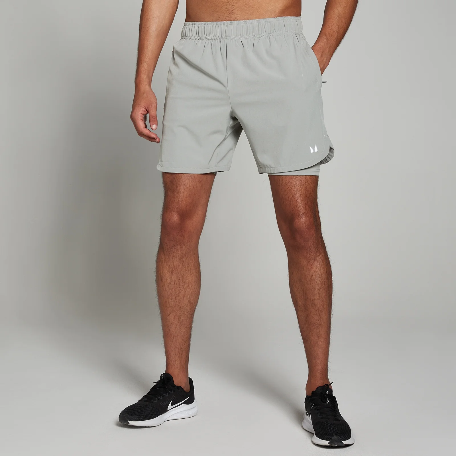 Een man draagt een stormgrijze MP 2-in-1 trainingsshort, met een elastische tailleband en een wit logo.
