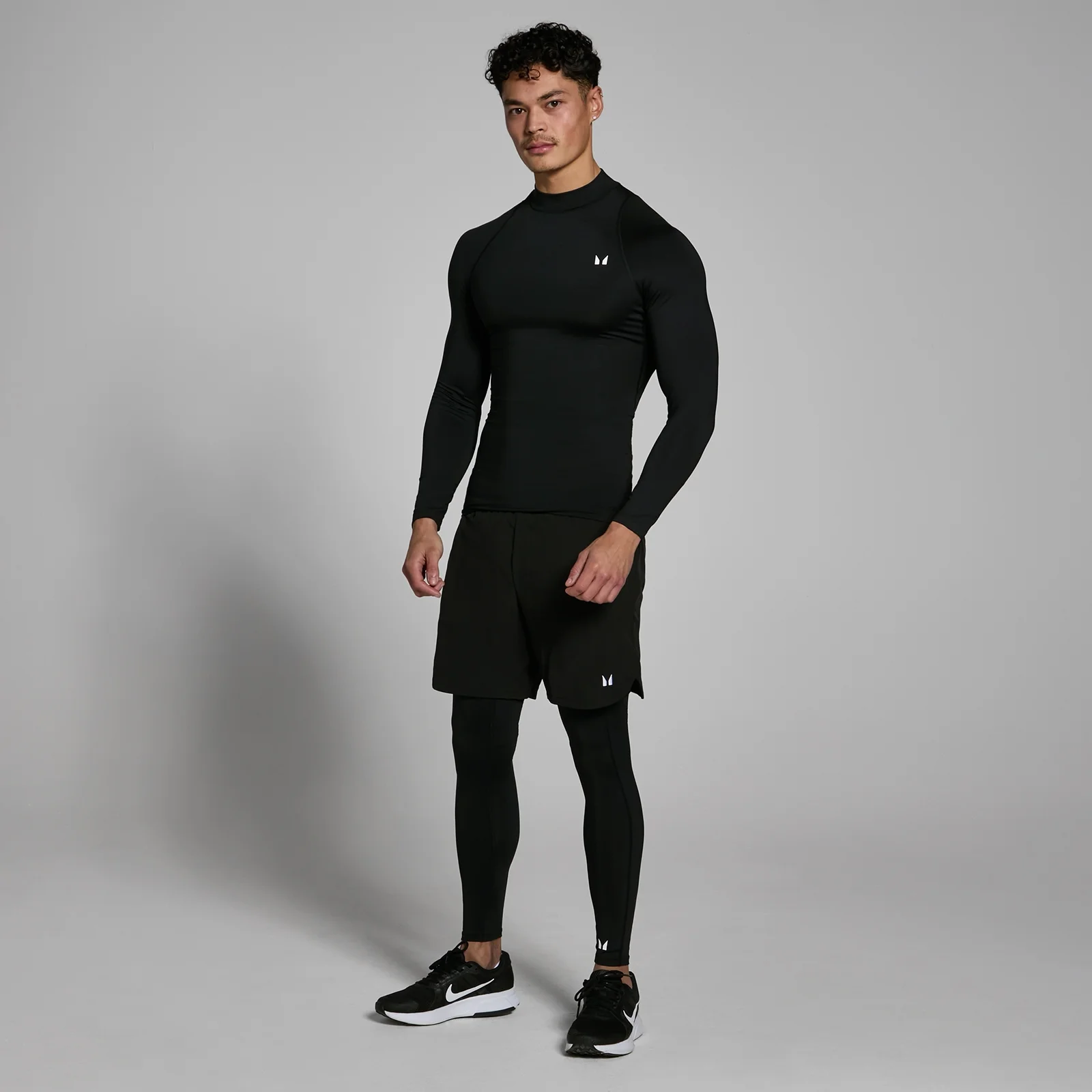 Een man draagt een zwarte, nauwsluitende top met lange mouwen, zwarte shorts en zwarte leggings.