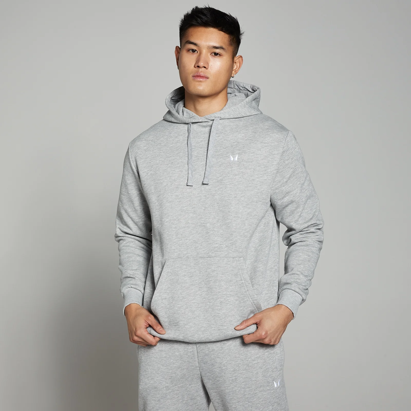 Een man draagt een gemêleerd grijze hoodie met capuchon en een klein wit logo.
