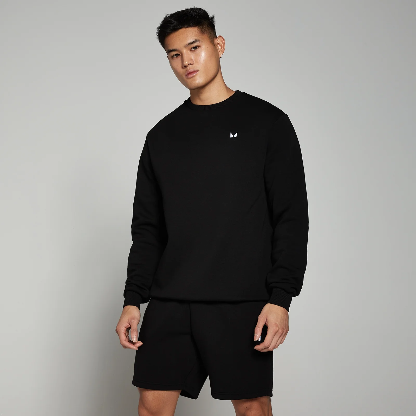 Een man draagt een zwarte sweatshirt met een klein wit logo en zwarte shorts.