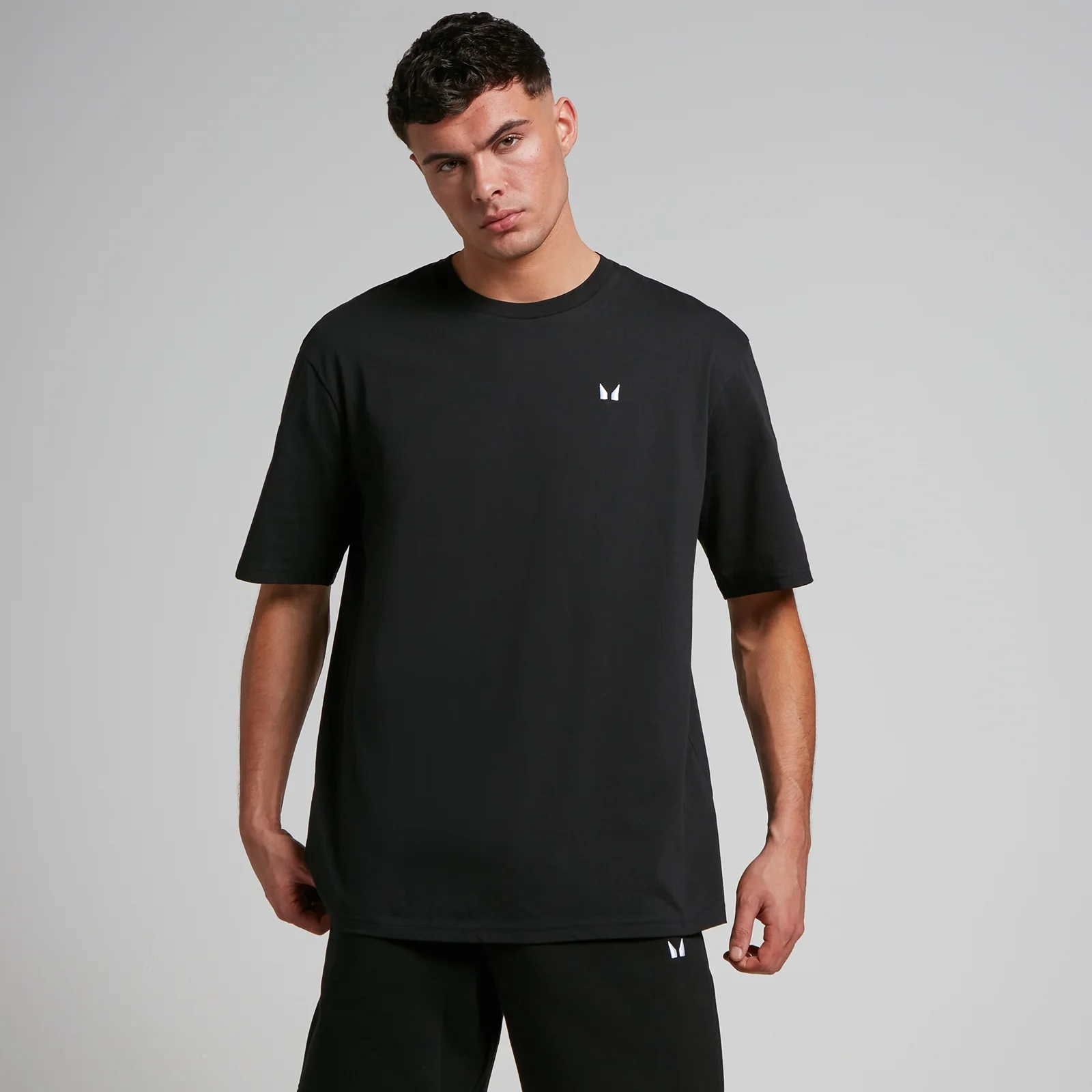 Een man draagt een zwart, oversized T-shirt met korte mouwen en een klein wit logo.