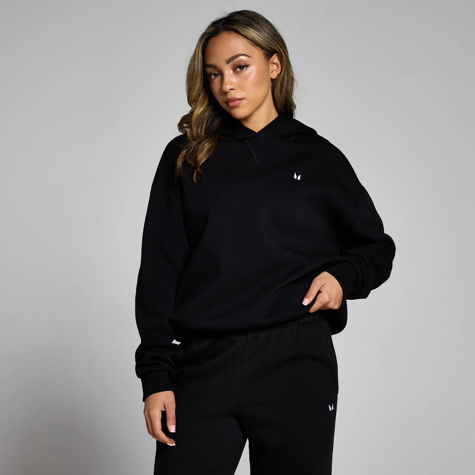 Een vrouw draagt een zwarte, oversized hoodie met een klein wit logo.