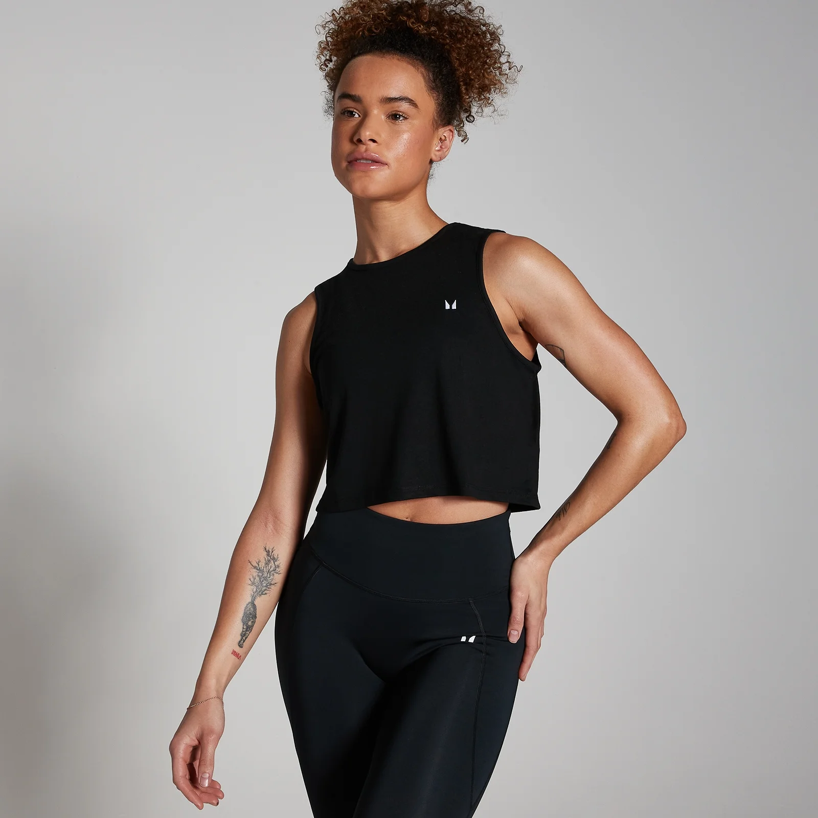 Een zwarte, mouwloze, korte sporttop met een klein wit logo, gedragen met een zwarte legging.