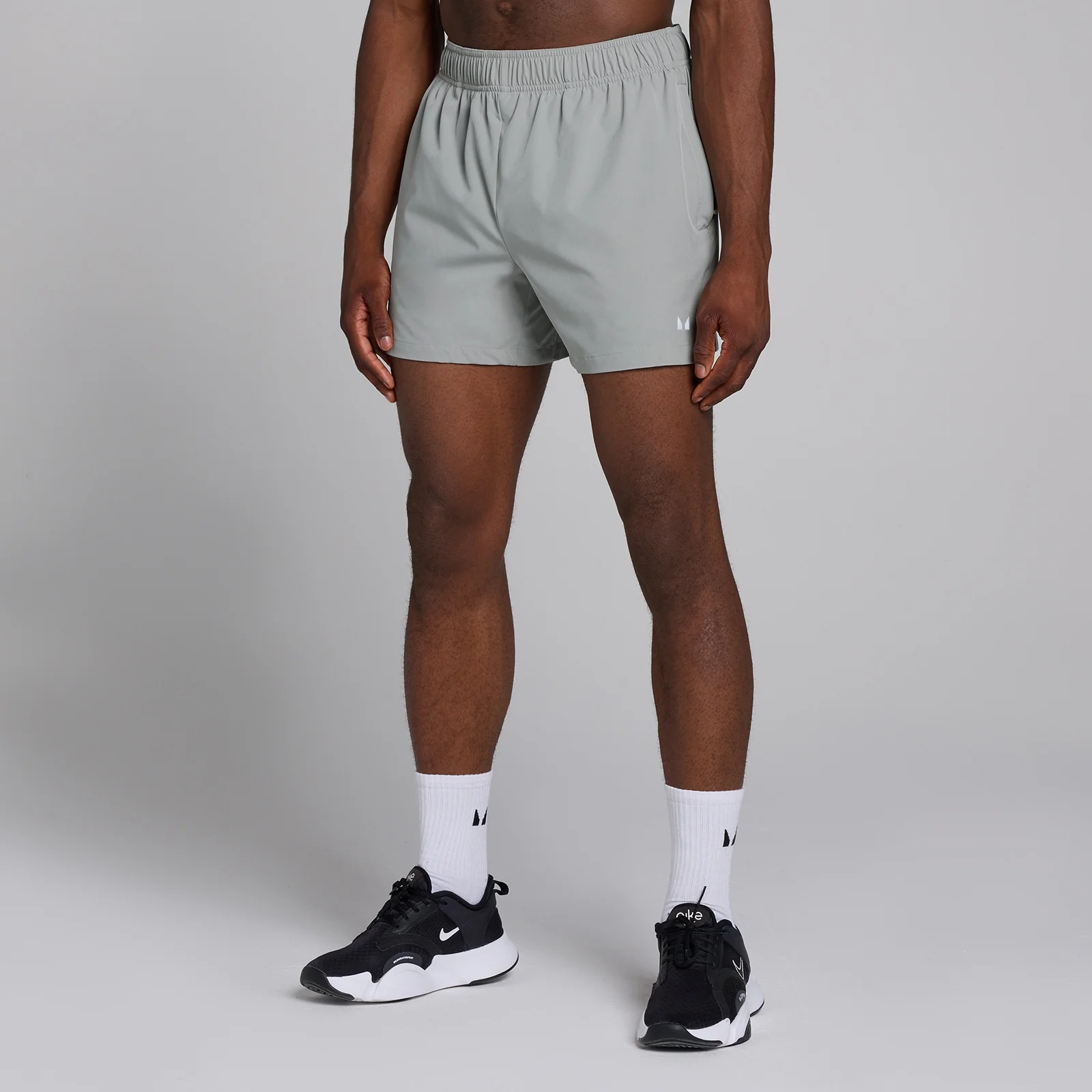 Een man draagt stormgrijze, geweven trainingsshorts met een elastische tailleband en witte sokken met zwarte sportschoenen.