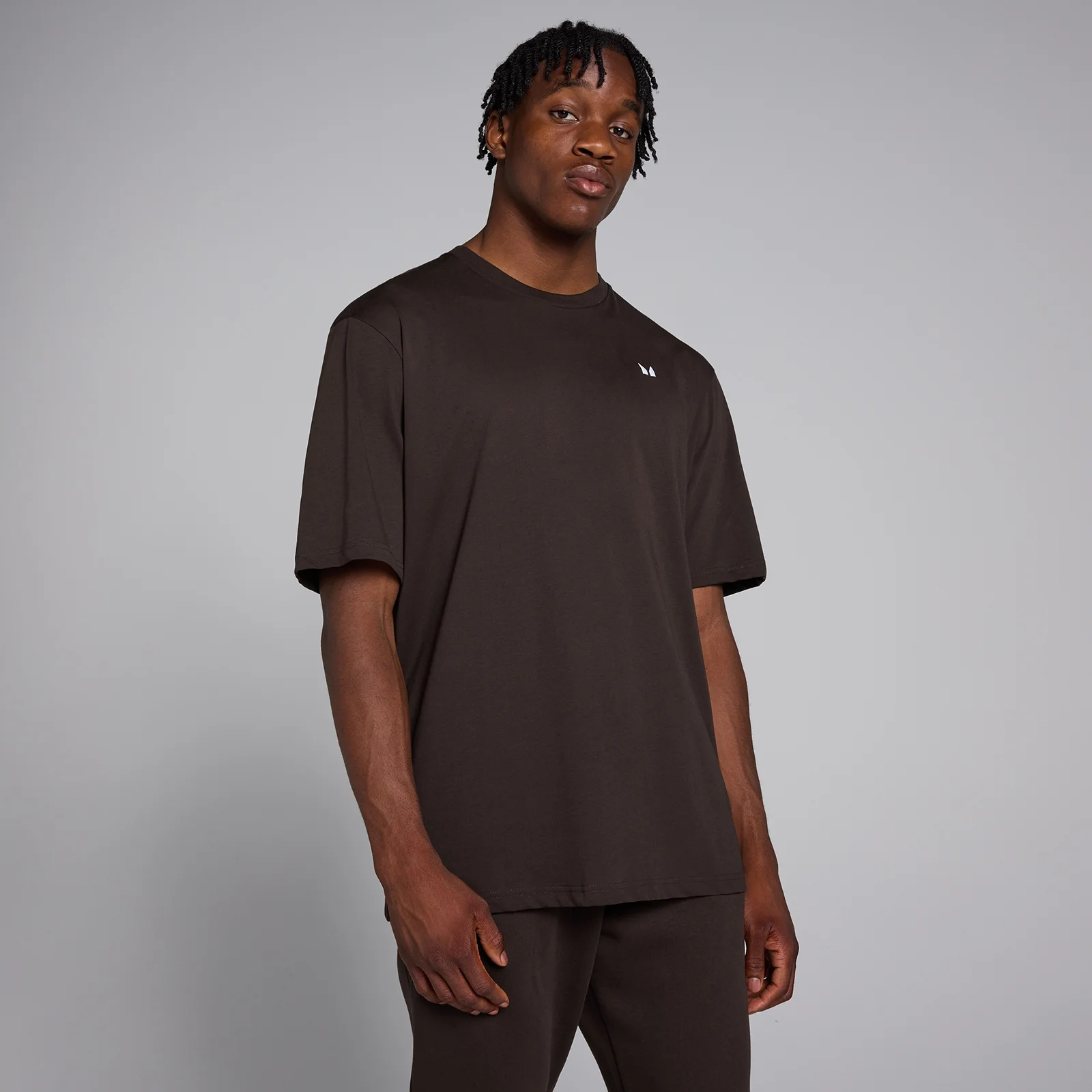 Een man draagt een koffiebruin, oversized T-shirt met korte mouwen en een klein wit logo.