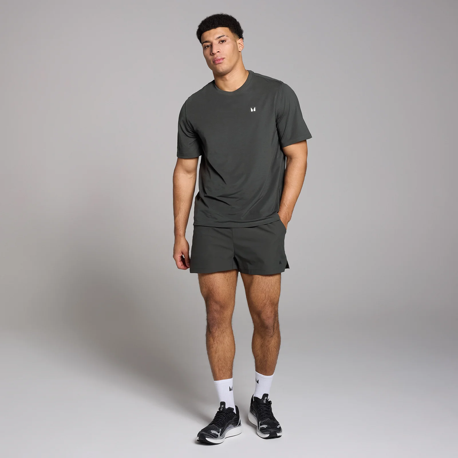 Een man draagt een zwart MP T-shirt en een zwarte korte broek, met witte sokken en zwarte schoenen.