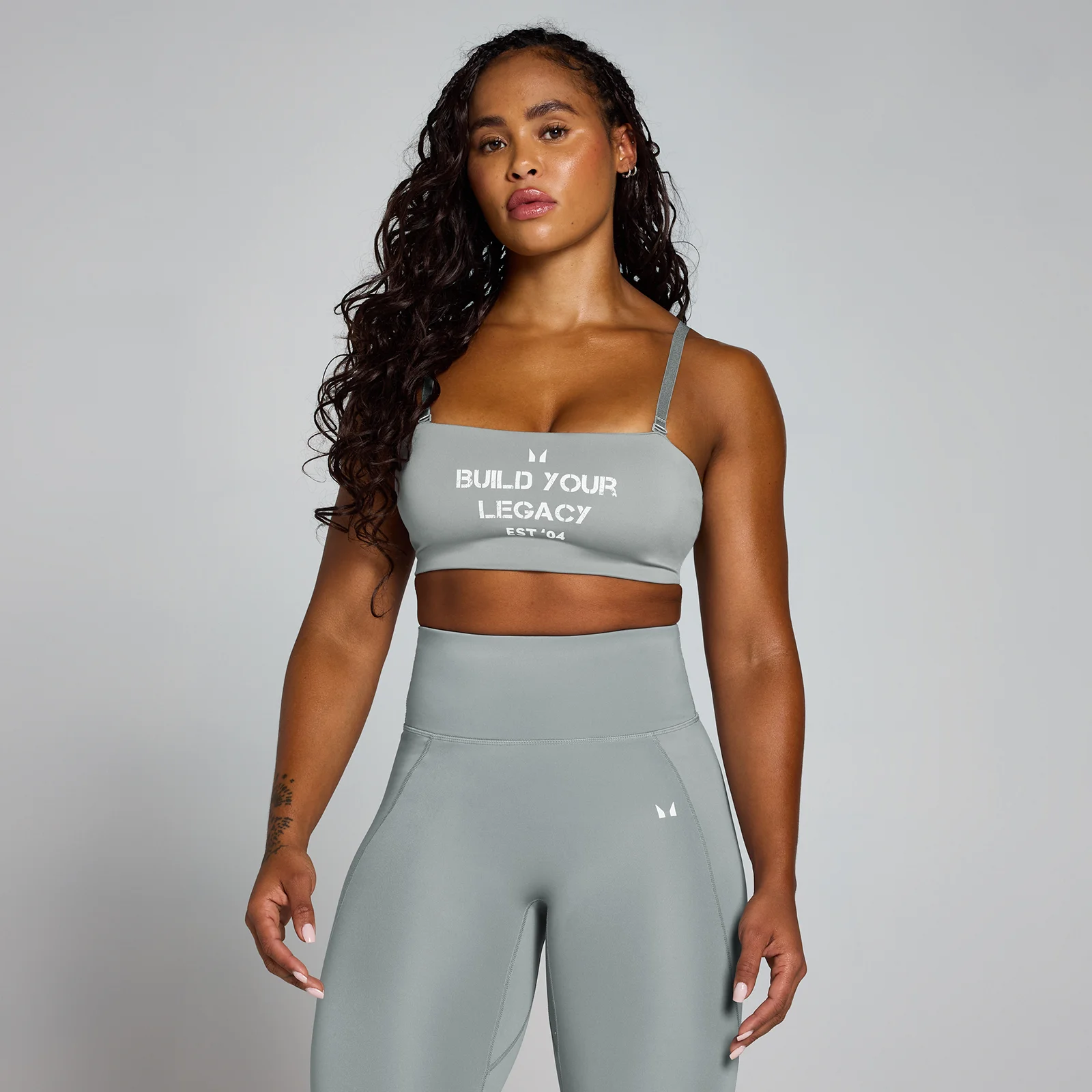 Een vrouw draagt een fossielgrijze sportbeha en legging met witte tekst "Build Your Legacy.