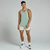 Een man draagt een vaalgroen hemd met witte accenten en beige shorts, staand tegen een grijze achtergrond.