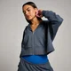 Een vrouw draagt een donkergrijze, korte hoodie met rits en een blauwe top.