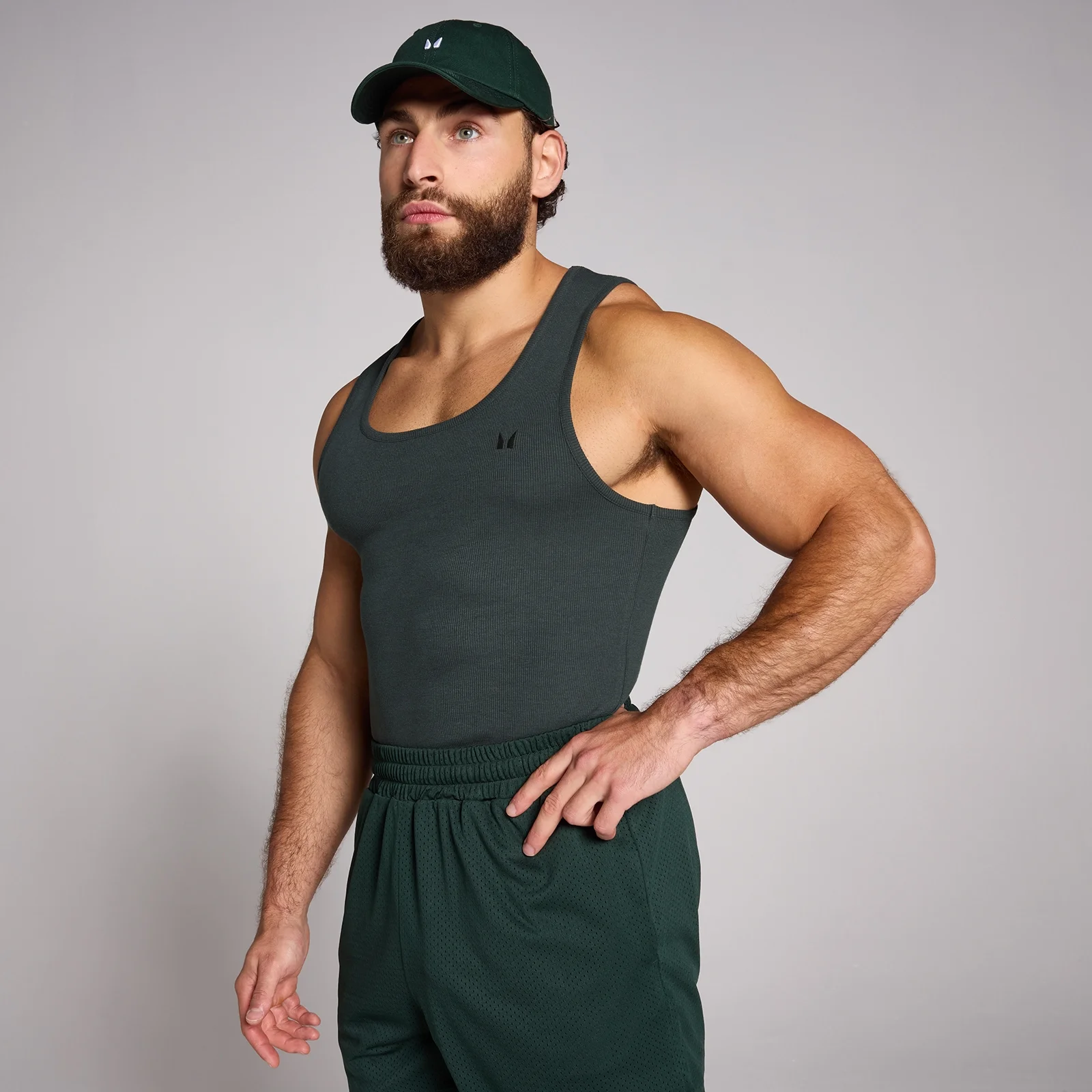 Een man draagt een mouwloze, geribbelde, donkergrijze tanktop en een groene pet en broek.