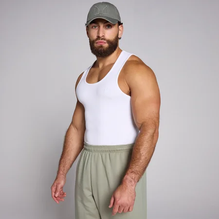 Een man draagt een witte, geribbelde tanktop en een groene pet en joggingbroek.