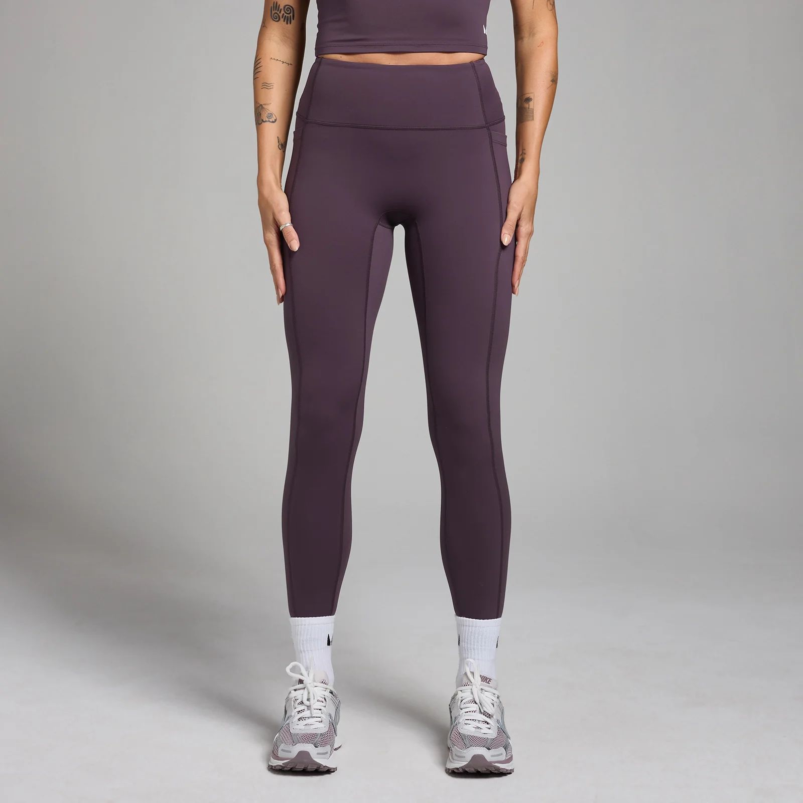 Een vrouw draagt een dieppaarse legging met zakken en sportschoenen.