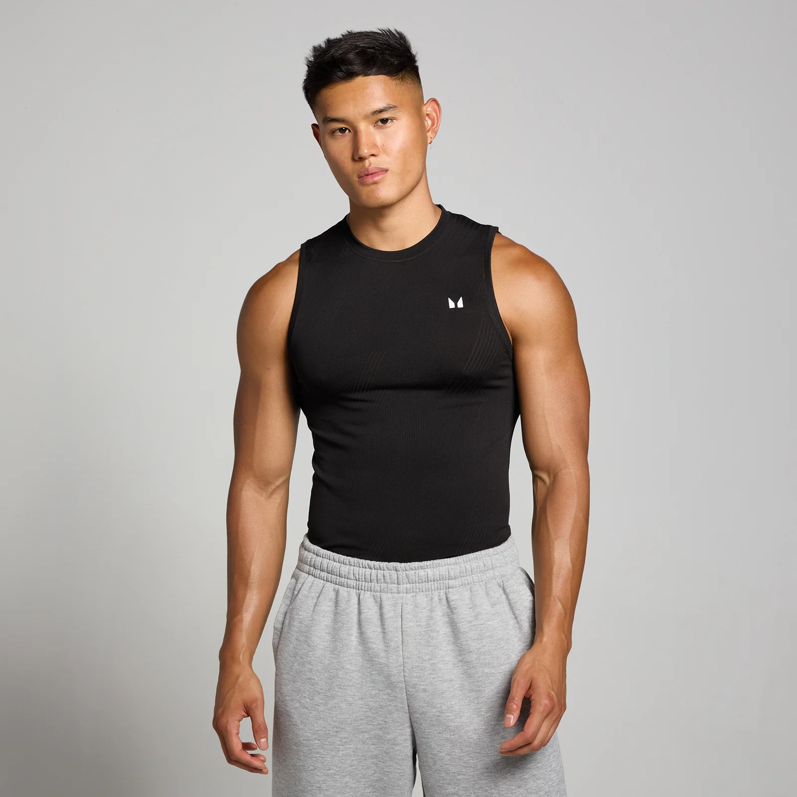 Een man draagt een zwart, mouwloos hemd met een klein wit logo, en grijze joggingbroek.