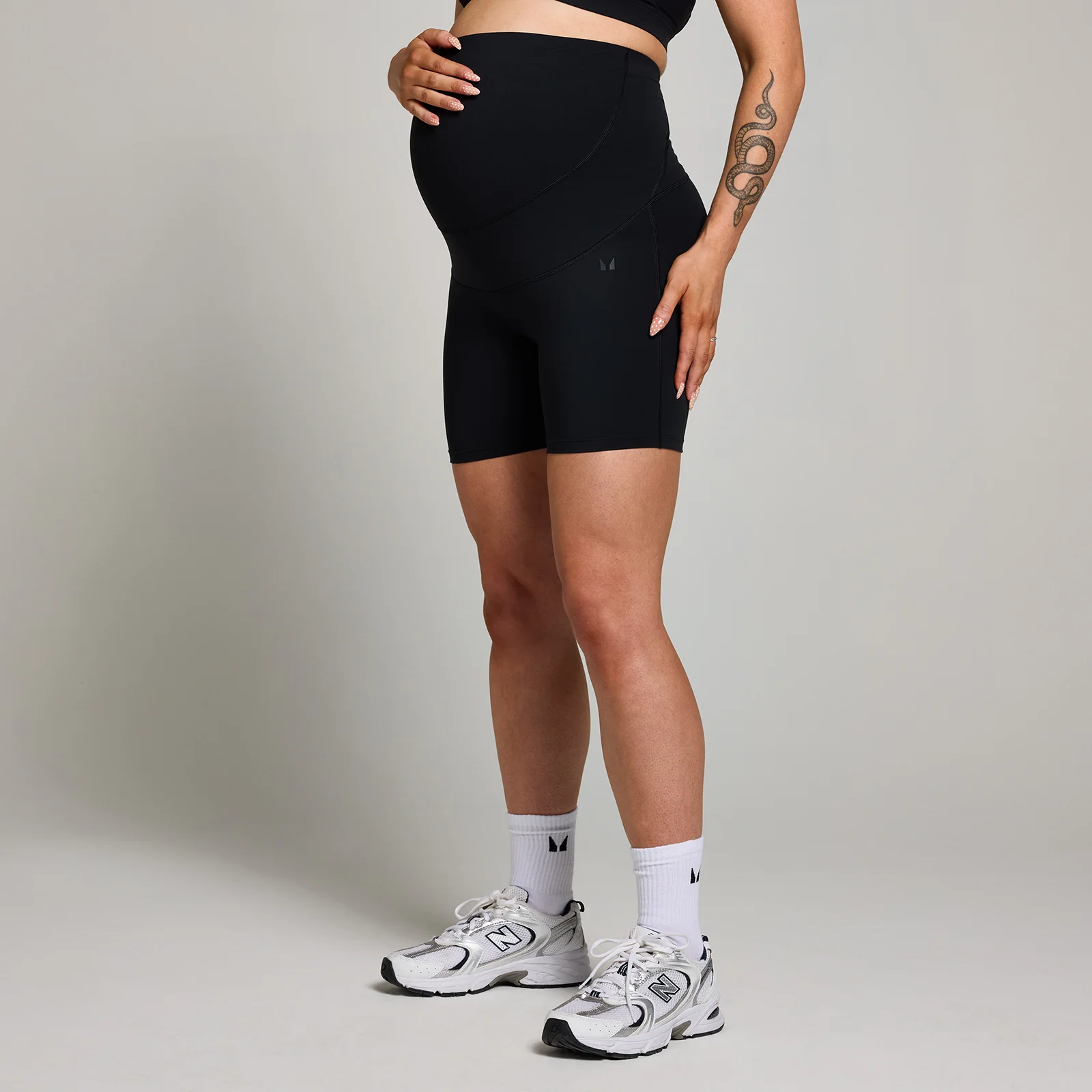 Een zwangere vrouw draagt een zwarte, korte sportbroek met hoge taille en witte sportschoenen.